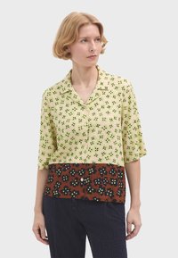 Korte mouwen shirt in lichtgroen met een bloemenpatroon, met een bruine sectie met donkere bloemen en knopen langs de knoopsluiting.