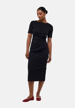 Femme portant une robe noire ajustée à manches courtes avec un détail froncé à la taille, associée à des chaussures plates rouges.