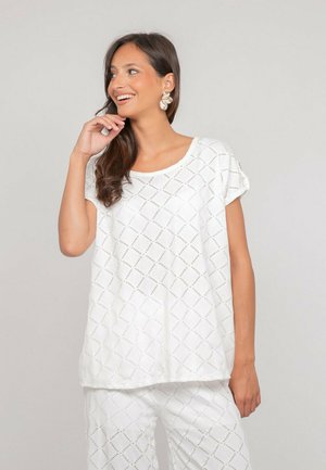 Femme souriante regardant vers la droite, vêtue d'un haut blanc à manches courtes et d'un pantalon assorti avec un motif perforé en forme de losanges, et portant de grandes boucles d'oreilles.