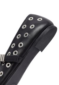 Scarpa di pelle nera con occhielli rotondi in argento, una punta ampia e un tacco basso. Presenta una fascia con fibbia per una maggiore sicurezza e una superficie superiore testurizzata.