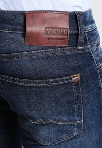 Mörkblå denimjeans med en läderpatch med Mustang-logotyp, synlig sömnad och detaljer i kopparfärg på bakfickan.