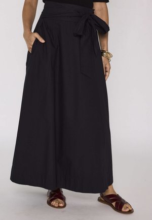 Jupe longue noire taille haute avec ceinture nouée, portée avec des bracelets dorés et des sandales en cuir marron foncé sur un fond clair.