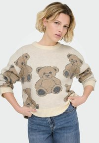 Beżowy sweter z brązowymi i szarymi wzorami misia, z prążkowanymi mankietami i dołem, luźny krój. Noszony na niebieskich dżinsach.