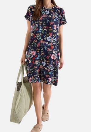 Femme en robe bleu marine à manches courtes avec imprimé floral multicolore, portant un grand sac cabas matelassé vert clair et des chaussures beiges sans lacets.