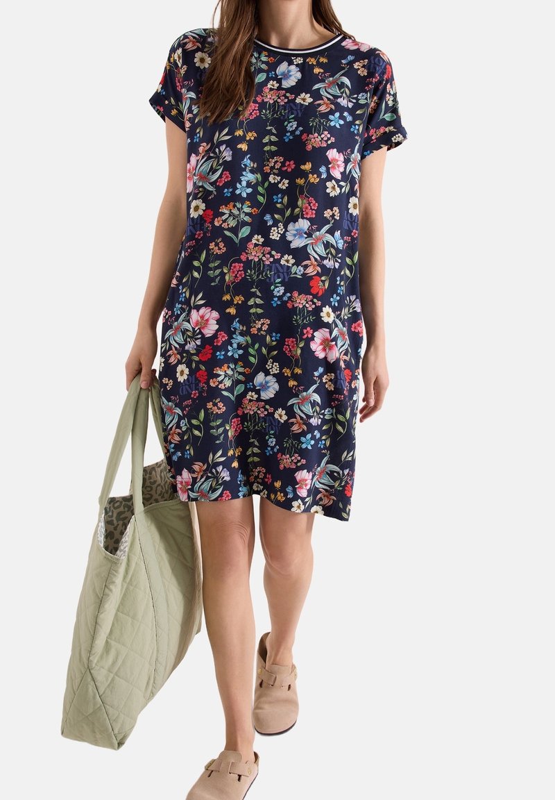 Femme en robe bleu marine à manches courtes avec imprimé floral multicolore, portant un grand sac cabas matelassé vert clair et des chaussures beiges sans lacets.