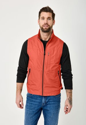 Mann mit Bart, der eine ärmellose rote Weste mit Reißverschluss über einem schwarzen Langarmshirt und blauen Jeans trägt, vor einem schlichten Hintergrund stehend.