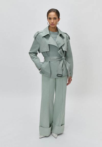 Γυναίκα που φοράει μαντό σε ανοιχτό πράσινο με ζώνη και παντελόνι με φαρδιά μπατζάκια, μαζί με λευκά μυτερά παπούτσια, στέκεται μπροστά σε απλό λευκό φόντο.