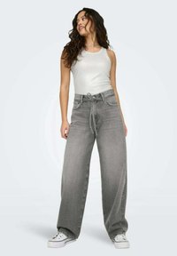 Jeans in denim grigio a gamba larga con vita regolabile, abbinati a una canotta bianca aderente e sneakers bianche su uno sfondo chiaro.