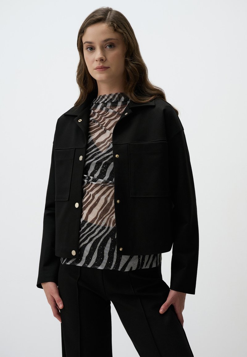 Zwart cropped jasje met gouden drukknoopjes, gecombineerd met een gelaagde top met een zebrapatroon in zwart- en grijstonen. Soepele textuur.