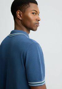Blaues Poloshirt mit einem gerippten Kragen, das grüne und weiße Streifen aufweist. Kurze Ärmel mit passenden gestreiften Akzenten. Weiche Textur.
