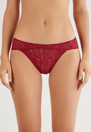 String rouge en dentelle avec des motifs floraux et des bords festonnés, présentant une texture douce et un design taille basse.