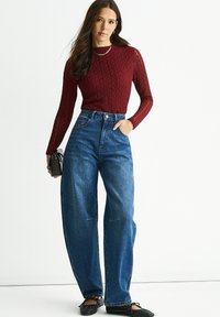 Red textured knit long-sleeve top paired with high-waisted, wide-leg blue denim jeans. Model is holding a small black bag.
