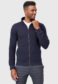 Mørkeblå zip-up jakke med tekstureret stof, ribbedeærmer og talje. Har en glat lynlås og et lille logo på brystet.