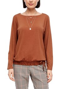 Brązowa bluzka z długim rękawem, zebrana na dolnej krawędzi i z detalem sznurka, zestawiona z długimi spodniami w kratę, w na przemian brązowych i kremowych kolorach.