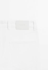 Jean en denim blanc avec une poche cousue et une étiquette en cuir rectangulaire lisse portant une marque embossée à la ceinture.