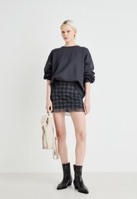Sweatshirt noir surdimensionné, jupe mini en tartan avec un ourlet festonné, bottines noires à la cheville, et portant un sac à dos de couleur claire.