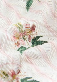 Tessuto bianco testurizzato con motivi geometrici in rilievo e fiori rosa stampati e foglie verdi in messa a fuoco soffusa.