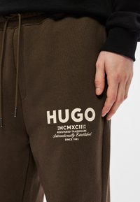 Καφέ φόρμες με λευκό λογότυπο και κείμενο με την επωνυμία "HUGO" και ημερομηνία ίδρυσης, κατασκευασμένες από μαλακό, υφαντό ύφασμα.