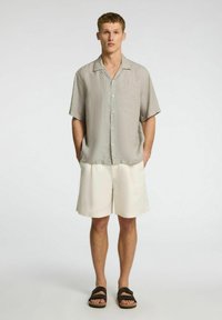 Koszula z krótkim rękawem w jasnoszarym kolorze, z zapięciem na guziki z przodu, zestawiona z kremowymi szortami. Stylizacja charakteryzuje się luźnym krojem i fakturowanym materiałem.