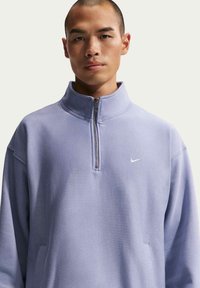 Sweatshirt à fermeture éclair violet clair avec col haut, texture douce et logo Nike blanc sur la poitrine gauche. Comprend des poches latérales.