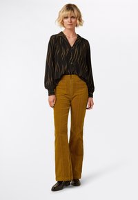 Blusa nera con motivo a linee ondulate dorate, abbinata a pantaloni in velluto a coste color senape a zampa d'elefante. Modella che indossa scarpe nere, in piedi davanti a uno sfondo neutro.
