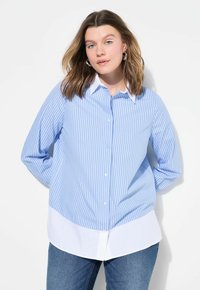 Camisa a rayas azul claro con cuello y dobladillo blancos, con mangas largas y botón en la parte delantera, llevada sobre unos pantalones vaqueros oscuros.