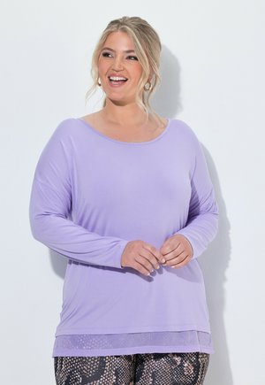 Lachende vrouw met blond haar in een lavendelgekleurde top met lange mouwen en leggings met patroon, staand voor een witte achtergrond.