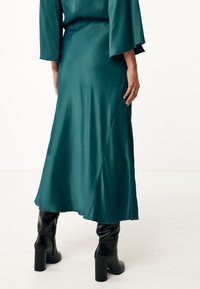 Sukienka maxi z satyny w kolorze teal, o płynnej linii, z wysokim stanem i delikatnym kształtem A-linii, zestawiona z czarnymi kozakami na obcasie do kolan.