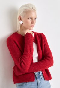 Cardigã de lã vermelha com decote redondo, fecho com botões e mangas ajustadas. Combinado com uma blusa branca com textura em relevo e jeans azul claro.