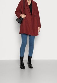 Röd texturerad kappa med slagkrage och två knappar, bärs över en svart topp och blå smala jeans, matchad med svarta ankelboots.