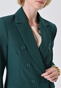Türkiser doppelt angeknöpfter Blazer mit sechs passenden Knöpfen, Reverskragen und glattem Stoff. Nahaufnahme, die ein beiges Oberteil darunter zeigt.