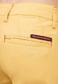 Gele katoenen broek met rode stiksels. Heeft een achterzak met een bordeauxkleurig label waarop "CIPO & BAXX" staat. Glad textuur, casual ontwerp.