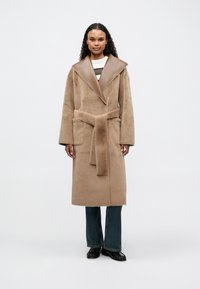 Rudas paltas su dideliu ruduo odiniu apykakle, diržu ir šoninėmis kišenėmis; minkštas, tekstūruotas audinys; nešiojamas su džinsais ir juodais batais.