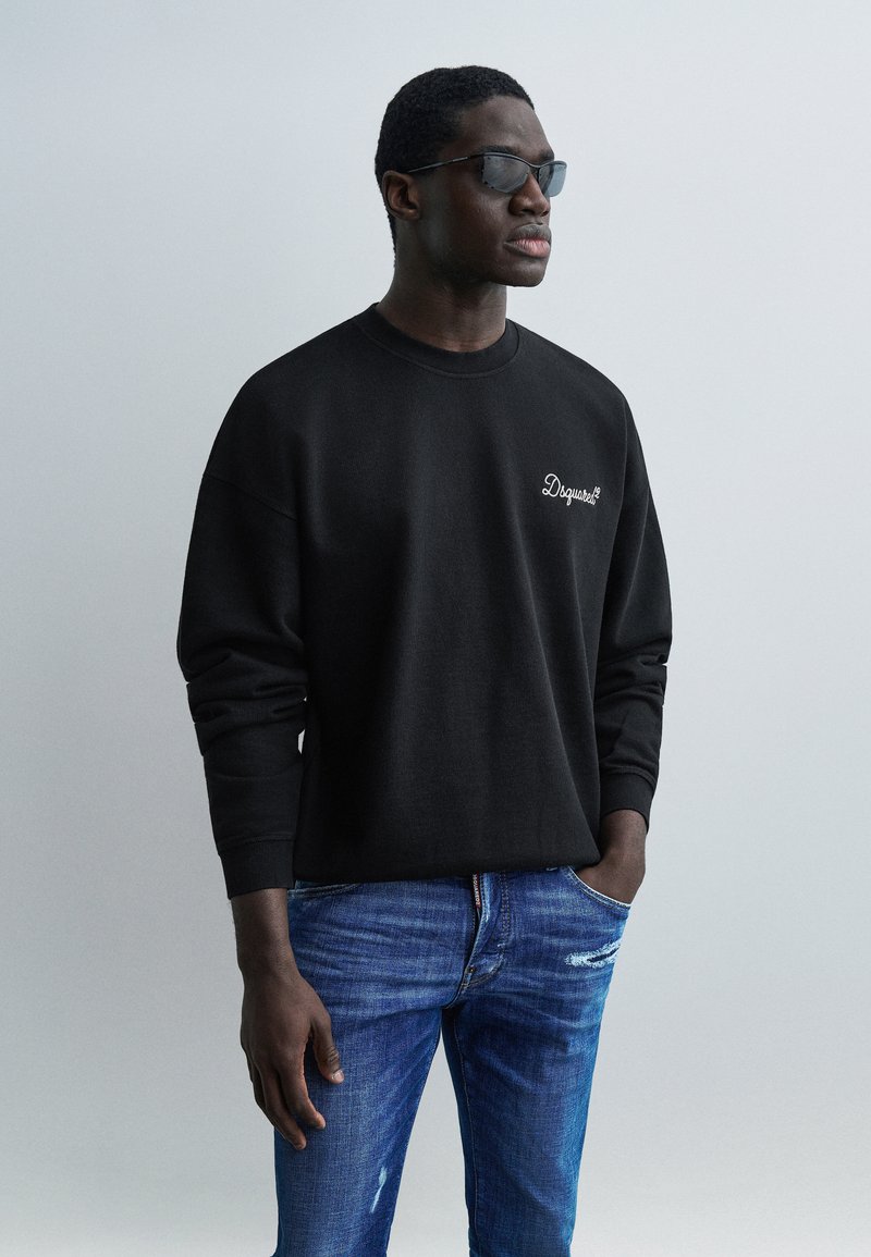 Άνδρας που φοράει μαύρο φούτερ με λευκό λογότυπο και μπλε τζιν, με τα χέρια στις τσέπες, κοιτάζοντας πλάγια, μπροστά σε απλό γκρι φόντο.