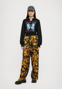 Sieviete, tērpusies melnā Versace Jeans Couture kapučjakā ar zilu tauriņu, dzeltenās baroka raksta biksēs, melnās zābakos un pelēkā čības cepurē, stāv taisni.