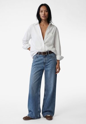 Kobieta ubraną w rozpiętą białą koszulę zapinaną na guziki, szerokie niebieskie dżinsy z brązowym paskiem i brązowe buty, stojąca na tle jednolitego białego tła.