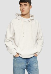 Sudadera de algodón beige claro con bolsillo tipo canguro, cordones ajustables y puños acanalados, combinada con jeans de mezclilla azul claro.