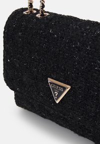 Svart texturerad handväska med en kviltad design, som har en gyllen triangel LOGO och kedjerem. Accentuerad med glittrande detaljer.