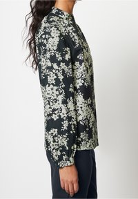 Blouse fleurie en noir et vert clair. Col haut, manches longues avec poignets élastiques, et coupe fluide. Tissu à motif.