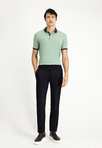 Polohemd in lichtgroen met marineblauwe kraag en mouwen, gecombineerd met donkerblauwe broek en donkerbruine instap schoenen, staand op een neutrale achtergrond.