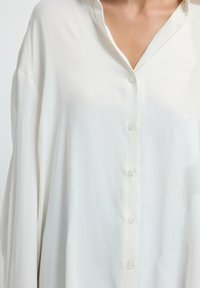 Chemise blanche à boutons, en tissu léger, avec un col, des manches longues et une texture lisse avec des boutons visibles sur le devant.