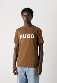 Vyras su trumpais drediukais, vilkintis rudą marškinėlį su užrašu "HUGO" baltais rašmenimis ir šviesiai mėlynomis džinsinėmis kelnių, stovi prieš paprastą foną.