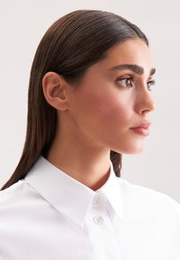 Camicia bianca con colletto e bottoni, tessuto liscio, caratterizzata da un design aderente. Capelli lunghi e lisci, con texture naturale.