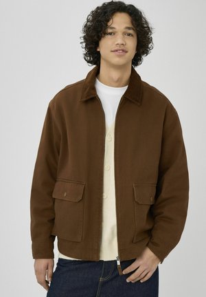 Veste en velours côtelé marron avec un col, deux poches frontales et une fermeture éclair en bas. Portée par-dessus un cardigan crème à boutons.