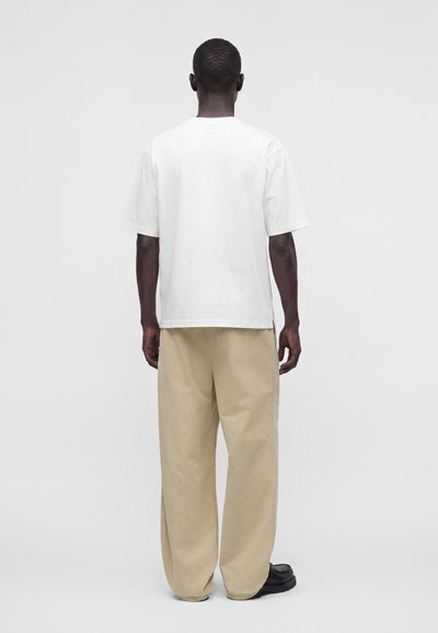 Άνδρας με κοντά μαλλιά που φοράει απλό λευκό κοντομάνικο μπλουζάκι, μπεζ φαρδιά παντελόνια και μαύρα παπούτσια, στέκεται με την πλάτη σε λευκό φόντο.