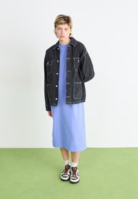 Črna denim jakna z belim šivom, oblečena čez svetlo modro obleko. Rjavi in beli športni copati dopolnjujejo obleko. Zeleno tla v ozadju.