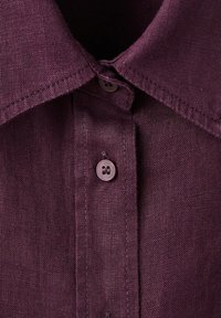Gros plan sur le col d'une chemise à boutons violette, avec la texture du tissu et deux boutons visibles sur la patte de boutonnage.