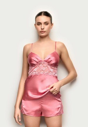 Młoda kobieta ubrana w różową satynową i koronkową kamizelkę oraz pasujące krótkie spodenki, stojąca na jasnym tle, trzymająca tkaninę na wysokości talii.