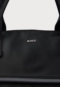 Mustakangas laukku mustilla hihnoilla, jossa on pieni hopeinen "BOSS"-logo keskittyneenä yläreunan lähelle.