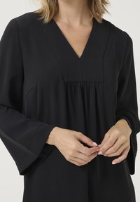 Blouse noire avec un col en V, des détails froncés et de longues manches cloche. En tissu lisse à finition mate.
